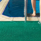 Close view of green ribbed barefoot matting at a pool edge, a swimmer stepping out of the water onto it beside the ladder rail