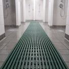 Green ribbed barefoot matting running between two rows of showers in a communal shower block, water draining through the ribs