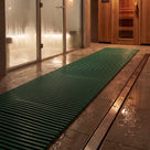 Green ribbed barefoot matting as a wet-side walkway between a glass steam room and timber sauna door, drain channel alongside