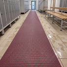 Mulberry red ribbed barefoot matting runner down a changing room aisle between lockers and timber benches, wet footprints on the tiles