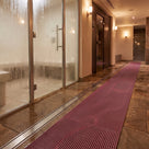 Mulberry red ribbed barefoot matting as a wet-side walkway between a glass steam room and timber sauna door, drain channel alongside