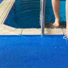 Close view of ocean blue ribbed barefoot matting at a pool edge, a swimmer stepping out of the water onto it beside the ladder rail