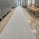 White ribbed barefoot matting runner down a changing room aisle between lockers and timber benches, wet footprints on the tiles