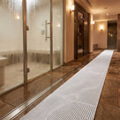 White ribbed barefoot matting as a wet-side walkway between a glass steam room and timber sauna door, drain channel alongside