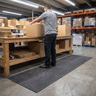 Dark grey 3mm ribbed PVC matting runner laid along a warehouse packing bench, worker standing on it taping a box