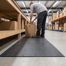 Dark grey 3mm ribbed PVC matting laid flat along a warehouse packing bench, worker standing on it taping a box