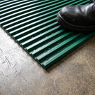 Close-up of green ribbed PVC matting edge showing the rib profile, water beads in the grooves and a safety boot