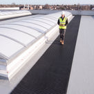 Black open-lattice matting beside barrel-vault rooflights on a light grey membrane roof, inspector walking it