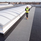 Light grey ribbed walkway matting beside barrel-vault rooflights on a dark roof, inspector carrying a clipboard