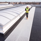 White ribbed walkway matting alongside barrel-vault rooflights on a dark roof, inspector in hi-vis walking it