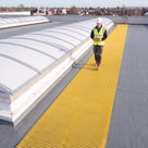 Yellow open-lattice matting beside barrel-vault rooflights on a grey membrane roof, inspector with a clipboard