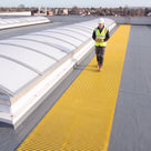 Yellow ribbed walkway matting beside curved rooflights on a dark grey membrane roof, inspector walking towards camera