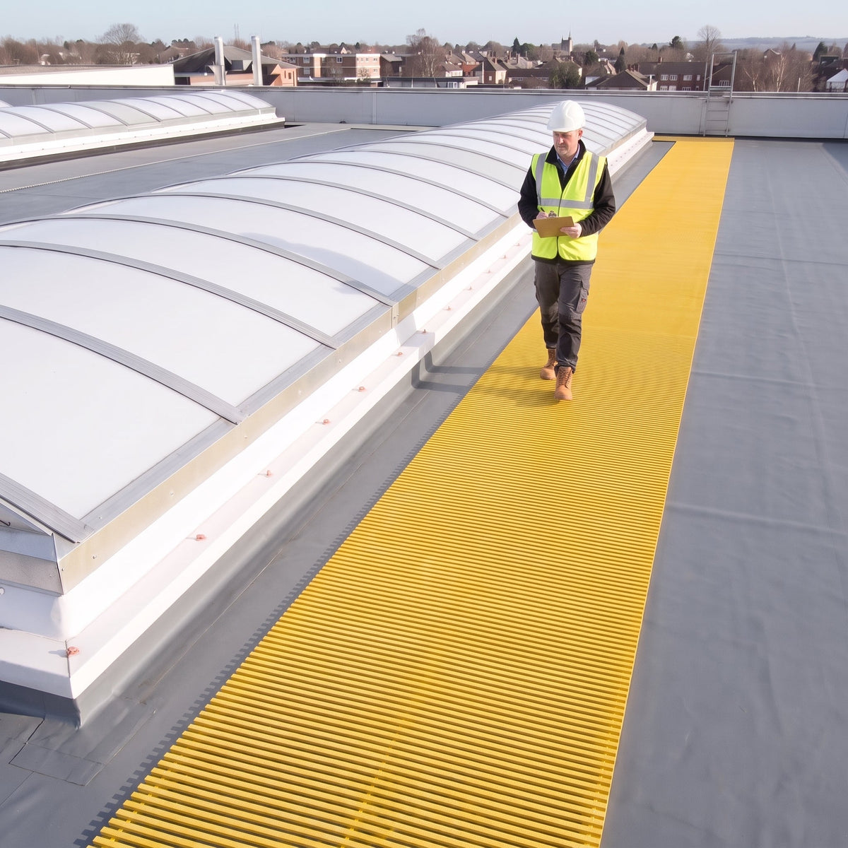 Yellow ribbed walkway matting beside curved rooflights on a dark grey membrane roof, inspector walking towards camera