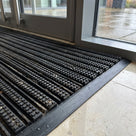Close view of the narrow scraper bars topped with square grip blocks on loose-laid black entrance matting, grit trapped in the channels