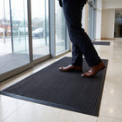Black solid-back scraper entrance mat laid loose on a polished office lobby floor inside the glass doors, a visitor stepping onto it