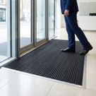 Black scraper entrance mat recessed flush in an aluminium-framed mat well inside the glass doors of an office lobby
