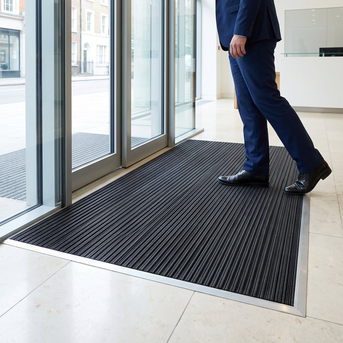 Black scraper entrance mat recessed flush in an aluminium-framed mat well inside the glass doors of an office lobby