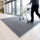 Grey solid-back scraper entrance mat laid loose inside a supermarket entrance, a shopper pushing a trolley across it
