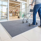 Grey scraper entrance mat recessed flush in a mat well at a supermarket entrance, a shopper pushing a trolley across it