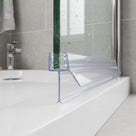 Clear seal along a wet glass bath screen on a white bath rim, chrome tap and grey tiles behind