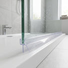 Clear seal on a green-edged glass bath screen, broad blade resting on the white bath rim, wet grey tiles behind