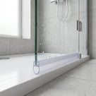 Clear bulb seal along the base of a wet glass shower door on a white tray, chrome hinges and hose behind