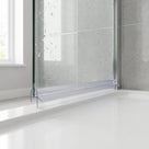 Clear seal along the bottom of a wet glass bath screen on a white bath rim, grey tiles and window behind