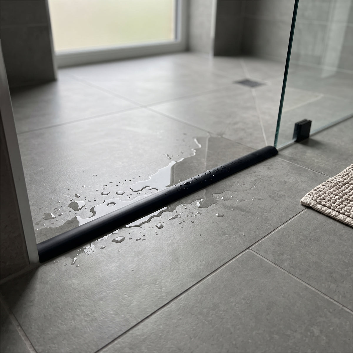 Slim black threshold bead across a grey tiled wet room floor, pooled water beside a glass panel