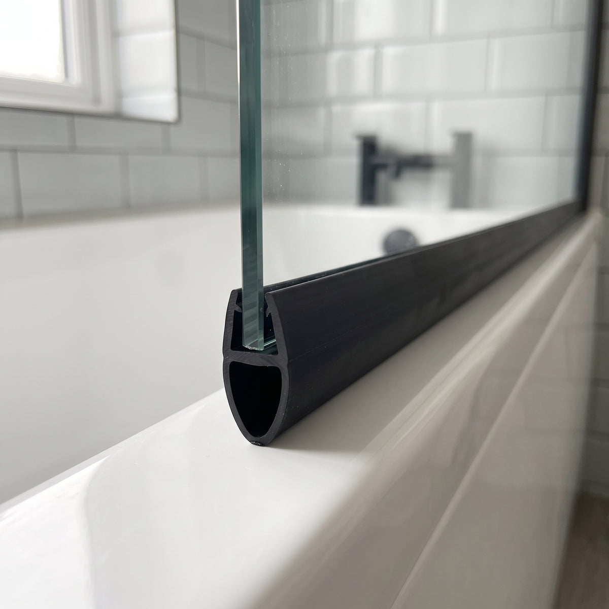 Black seal with a hollow bulb on a glass bath screen edge, white metro tiles and black tap behind