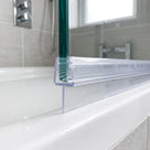 Clear seal on the bottom edge of thick glass over a white bath, chrome valve and grey tiles behind