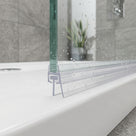 Clear carrier and T seal along the bottom of a glass shower panel on a white tray, grey tiles and a plant behind