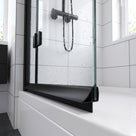 Black deflector seal along a bath screen bottom edge, wet glass and white tiles behind