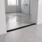 Black threshold strip laid across a wet room entrance, wet grey tiles and a glass panel behind it
