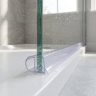 Clear seal along the base of a wet glass bath screen on a white bath, grey tiled bathroom and window behind