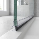 Clear seal on a glass shower screen edge beside a black deflector strip on a white tray, grey tiles