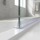 Clear seal at the foot of a green-edged glass panel on a white tray, sunlit grey tiles and towel rail behind