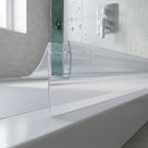 Clear seal gripping a green-edged bath screen, long blade sweeping onto the white rim, chrome valve behind