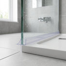 Clear seal along the base of a wet glass screen, blade reaching down past a white tray to grey floor tiles