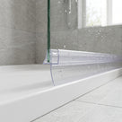 Clear seal along the bottom of a wet glass bath screen resting on a white bath rim, grey tiles behind