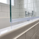 Clear tapered seal along a glass bath screen edge on a white bath rim, white metro tiles behind