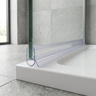 Clear bulb-loop seal on a glass screen edge meeting a white bath rim, concrete-look tiles, window left