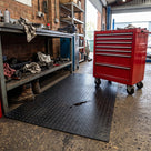 Black small-dot PVC matting in front of a garage workbench, a red tool chest on castors and an oil drip sitting on the surface