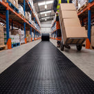 Black small-dot PVC matting unrolled from a roll as one continuous runner down a warehouse aisle, a worker pushing a sack truck of boxes beside it