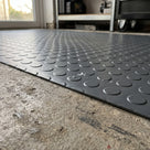 Low close view of the thin 1.6mm cut edge of dark grey small-dot PVC matting on concrete, water beading on the raised dots