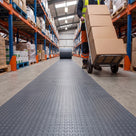 Dark grey small-dot PVC matting unrolled from a roll as one continuous runner down a warehouse aisle, a worker pushing a sack truck of boxes beside it