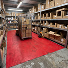 Red small-dot PVC matting on a stockroom floor between shelving, a roll cage and pallet truck standing on it