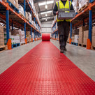 Red small-dot PVC matting unrolled as one continuous runner marking a pedestrian walkway down a warehouse aisle, a picker in hi-vis carrying a tote alongside