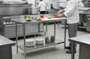 Chef preparing fresh vegetables on a 1200 x 600mm stainless steel prep table in a busy commercial kitchen, with mixing bowl, plates and storage trays on the galvanised undershelf