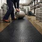 Black penny-stud PVC matting runner down a pub beer cellar, a cellarman rolling a steel keg onto it past beer drips