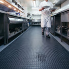 Dark grey penny-stud PVC matting runner down a restaurant kitchen walkway, a small spill held among the studs as a chef carries plates